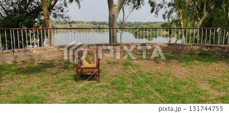 A chair on the grass near a railing overlooking a lake surrounded by trees and greenery A chair on the grass near a railing overlooking a lake surrounded by trees and greenery 131744755