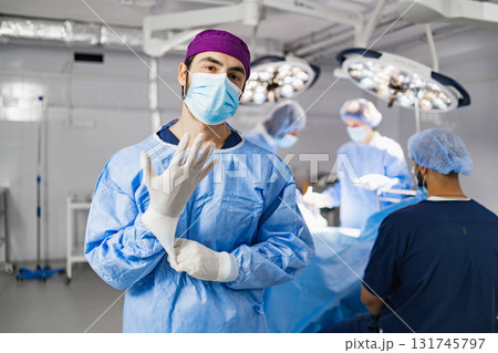 A surgeon wearing a mask and gloves in an operating room, preparing for a procedure with his team in the background. 131745797