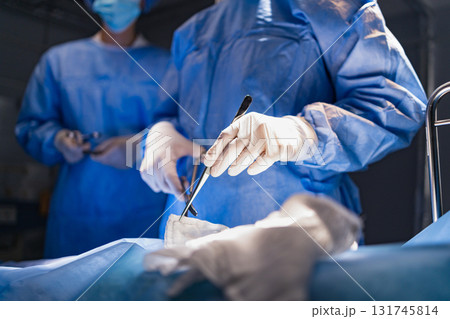 A surgeon uses a scalpel during an operation in a sterile operating room. Another medical professional assists in the background. A surgeon uses a scalpel during an operation in a sterile operating room. Another medical professional assists in the background. 131745814