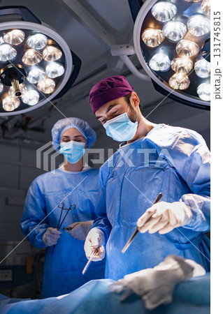 A surgeon and assistant perform surgery in a modern operating room, illuminated by bright surgical lights. 131745815