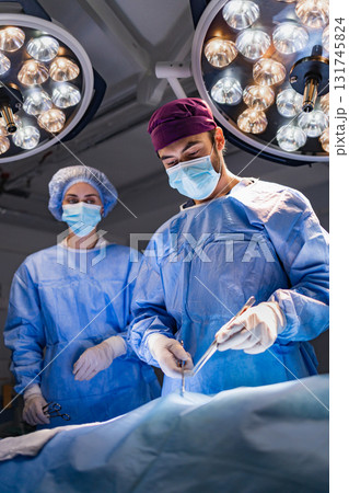A surgeon and assistant perform a procedure in a modern operating room, illuminated by bright surgical lights. 131745824