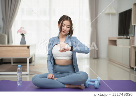 Young woman in activewear checking fitness tracker while sitting on exercise mat in modern living room with dumbbells and water bottle nearby Young woman in activewear checking fitness tracker while sitting on exercise mat in modern living room with dumbbells and water bottle nearby 131749050