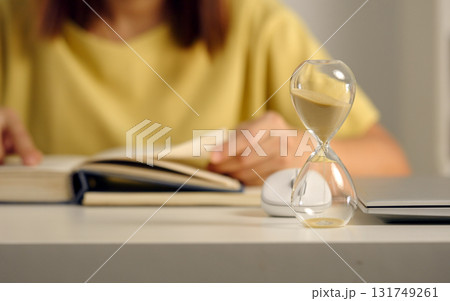Glass hourglass standing against background of woman reading a book closeup. Glass hourglass standing against background of woman reading a book closeup. 131749261