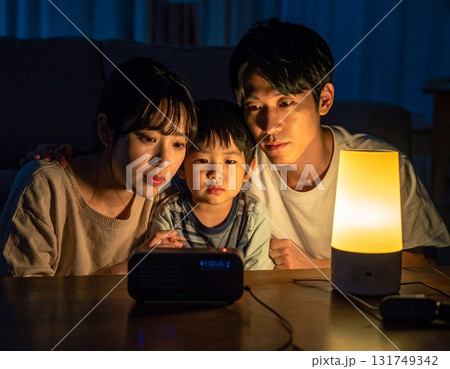 停電の夜、暗いリビングでラジオに耳を傾ける不安な表情の家族3人 131749342