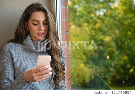 young sensual long-haired brunette stands by the window and uses a smartphone 131749894