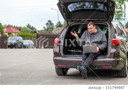 a man sitting in the open trunk of a car and working with a laptop, mobile technology, remote work 131749987