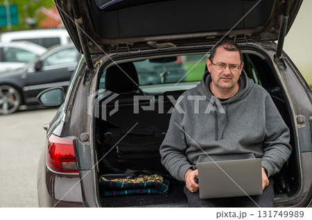 a man sitting in the open trunk of a car and working with a laptop, mobile technology, remote work 131749989