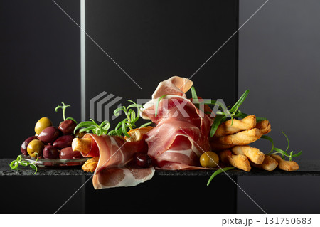 Prosciutto with bread sticks, olives, and rosemary on a black background. Prosciutto with bread sticks, olives, and rosemary on a black background. 131750683