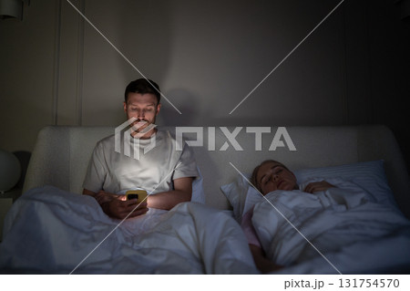 Tired man using smartphone lying in bed at night while wife sleeps beside him in dark bedroom Tired man using smartphone lying in bed at night while wife sleeps beside him in dark bedroom 131754570
