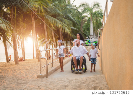 Disabled man in a wheelchair moves on a ramp to the beach with his family. Disabled man in a wheelchair moves on a ramp to the beach with his family. 131755613