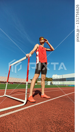 Confident male athlete resting near hurdle on running track under blue sky 131756826