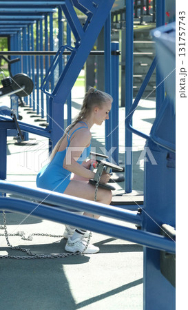 In a blue sports bodysuit, a woman lifts dumbbells at an outdoor gym, focusing on strength training exercises. The photo emphasizes the advantages of combining weight training with outdoor activities 131757743