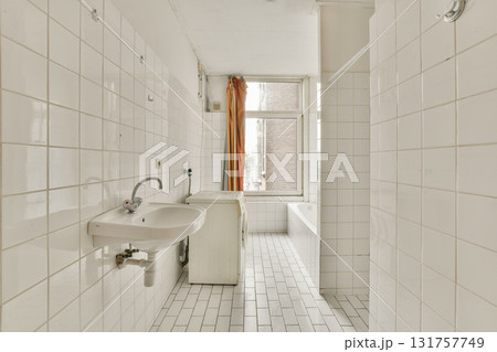 A spacious, minimalist bathroom featuring white tiles, a simple sink, and a freestanding bathtub illuminated by natural light from a window. 131757749
