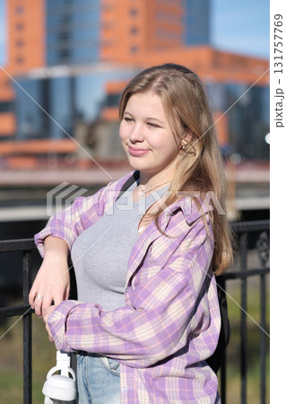 A teenage girl enjoys the sunlight as she poses by a railing, holding headphones in her hand. The photo captures the vibrancy and freedom of youth in an urban setting. 131757769