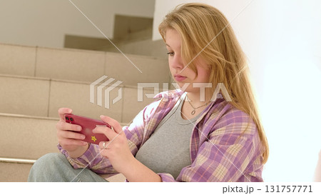 A young woman sitting on stairs smiles while looking at her phone. The image reflects casual moments of digital connection and the joy of staying in touch with loved ones. 131757771