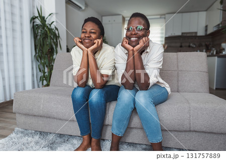 Two cheerful black twin sisters watching television sitting on sofa at home 131757889