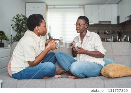 Two Young Adult Black Women Relaxing on Sofa, Drinking Tea and Chatting Two Young Adult Black Women Relaxing on Sofa, Drinking Tea and Chatting 131757890