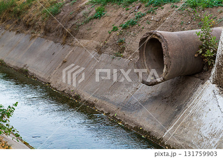 The end of an old concrete pipe of large diameter protrudes from the ground above a ditch with a flowing small river with reinforced concreted banks. Road underground communications The end of an old concrete pipe of large diameter protrudes from the ground above a ditch with a flowing small river with reinforced concreted banks. Road underground communications 131759903