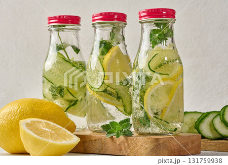 Glass bottles with red lids, filled with detox water containing cucumber and lemon slices and mint leaves, white table 131759938