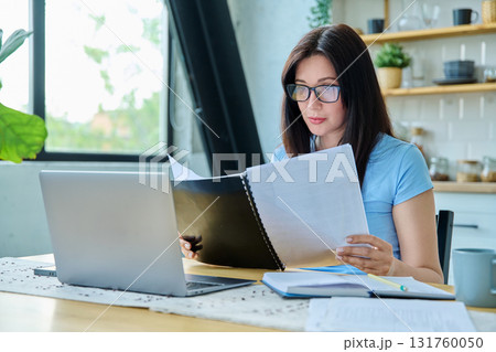 Young serious woman working with laptop, reading business papers, sitting at table at home 131760050