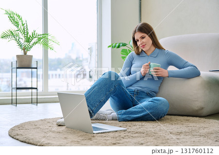Young relaxed woman with cup using laptop, sitting on floor at home Young relaxed woman with cup using laptop, sitting on floor at home 131760112