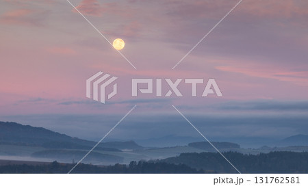 Full moon glows above a misty landscape, casting a soft, ethereal light over the scene. 131762581