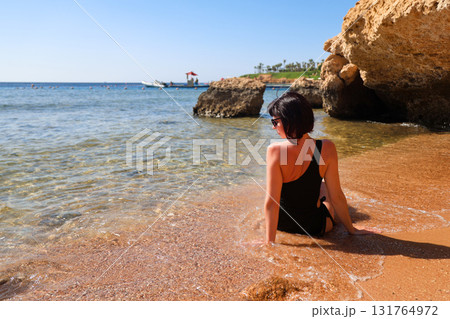 A brunette woman with glasses in a black bathing suit sits on the sand overlooking the sea, view from the back 131764972