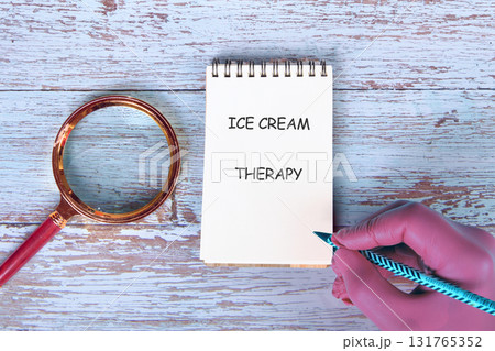 An open notebook sits on a table saying ICE CREAM THERAPY. A hand holds a pencil near the notebook, and a magnifying glass rests beside it, all on light blue wood 131765352