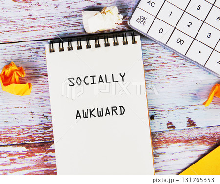 A notebook with the words SOCIALLY AWKWARD sits on a desk beside a calculator and some crumpled paper. The desk has a vintage wooden pattern 131765353