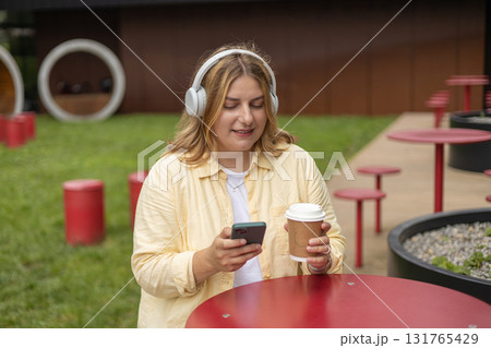 Happy young Caucasian woman in good mood listening to music with headphones and sitting on terrace street cafe in the city. Music lover enjoying music. Urban lifestyle concept. Happy young Caucasian woman in good mood listening to music with headphones and sitting on terrace street cafe in the city. Music lover enjoying music. Urban lifestyle concept. 131765429