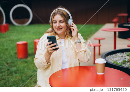 Happy young Caucasian woman in good mood listening to music with headphones and sitting on terrace street cafe in the city. Music lover enjoying music. Urban lifestyle concept. Happy young Caucasian woman in good mood listening to music with headphones and sitting on terrace street cafe in the city. Music lover enjoying music. Urban lifestyle concept. 131765430