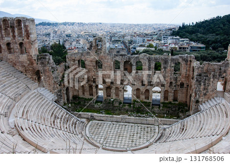A large open amphitheater with a stage in the center. The stage is surrounded by rows of empty seats. The amphitheater is located in a city with a view of the city below, the Acropolis of Athens. 131768506