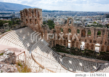 A large open amphitheater with a stage in the center. The stage is surrounded by rows of empty seats. The amphitheater is located in a city with a view of the city below, the Acropolis of Athens. 131768521