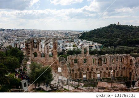 A large open amphitheater with a stage in the center. The stage is surrounded by rows of empty seats. The amphitheater is located in a city with a view of the city below, the Acropolis of Athens. A large open amphitheater with a stage in the center. The stage is surrounded by rows of empty seats. The amphitheater is located in a city with a view of the city below, the Acropolis of Athens. 131768533