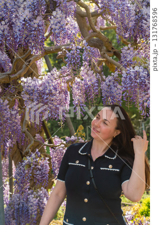 Young beautiful woman standing gracefully beside cascading purple wisteria flowers in bloom. 131768596