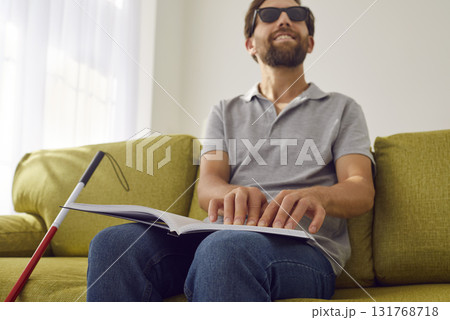 Blind smiling man sitting on sofa at home and reading tactile braille book. 131768718