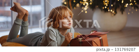 Banner smiling girl in pajamas holding gift box lying on cushion near Christmas tree with lights 131769207