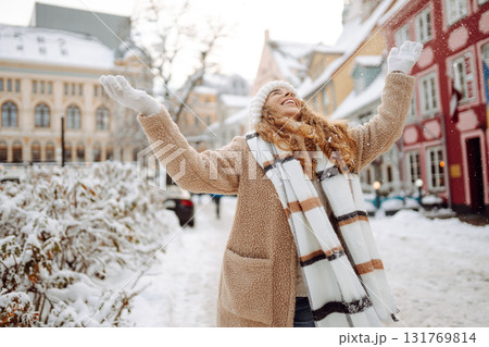 Young happy woman in a winter coat and scarf walks along a snowy city street. Concept of style. Young happy woman in a winter coat and scarf walks along a snowy city street. Concept of style. 131769814
