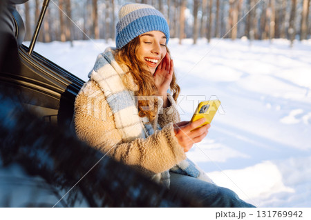 Happy Woman sitting in trunk, using phone. Snowy weather. Concept of adventure, travel by car. Happy Woman sitting in trunk, using phone. Snowy weather. Concept of adventure, travel by car. 131769942