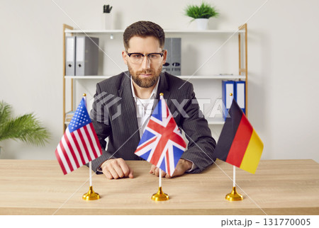 Pensive young man sitting at desk in office looking at flags of different countries 131770005