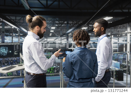 African american professionals handling performance metrics on tablet, ensuring technical workflow in server room. Employees team reviewing diagnostics for data center stability. African american professionals handling performance metrics on tablet, ensuring technical workflow in server room. Employees team reviewing diagnostics for data center stability. 131771244
