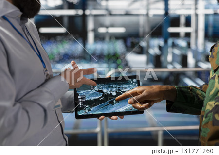 Military technician reviewing tactical dashboard on tablet screen in server hub, standing on industrial platform. Army expert using strategic communications and infrastructure software systems. 131771260
