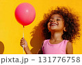 Child with big smile enjoys sunny day while holding a pink balloon against yellow backdrop 131776758