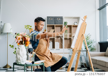 Asian male painter do artwork in art workshop, painting supplies, oil pastels, two canvas easel, creative space with paintbrush in art studio 131776807