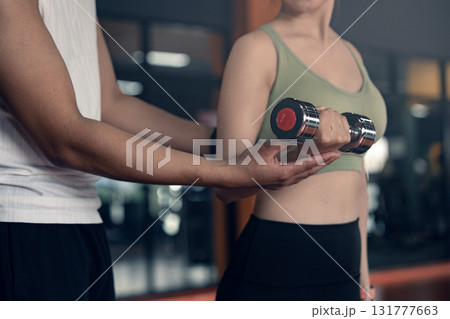 Young asian woman doing exercises with dumbbells in a gym while her trainer is supporting her. 131777663