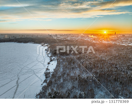 Snow-covered forest on lake shore with ice at sunset and the city on horizon, auerial view Snow-covered forest on lake shore with ice at sunset and the city on horizon, auerial view 131778369