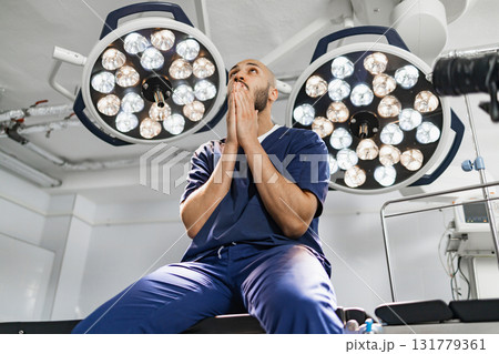 A surgeon in scrubs looks up with hands clasped in an operating room, illuminated by surgical lights. 131779361
