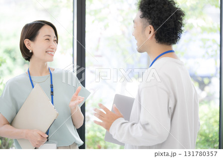 緑のある窓際で話す名札を付けた笑顔の男女のパソコンを持つ爽やか、話すオフィスカジュアルミドルビジネス 緑のある窓際で話す名札を付けた笑顔の男女のパソコンを持つ爽やか、話すオフィスカジュアルミドルビジネス 131780357