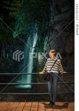 Woman enjoys a quiet moment by a waterfall in a lush, natural setting during the evening hours Woman enjoys a quiet moment by a waterfall in a lush, natural setting during the evening hours 131787143