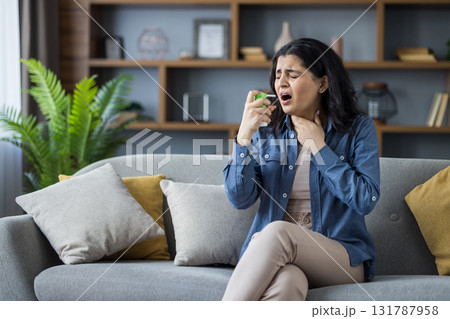 Young Indian woman sitting on sofa at home, holding her neck with her hand, feeling severe pain, using medical spray. 131787958